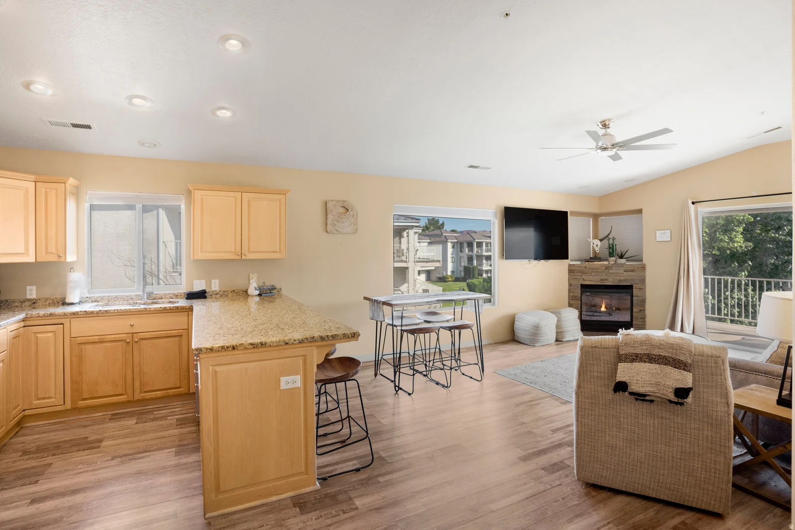Spacious living area featuring wood-finish flooring, a kitchen with light wood cabinetry and granite-style countertops, a built-in fireplace with stone surround, recessed lighting, and a ceiling fan