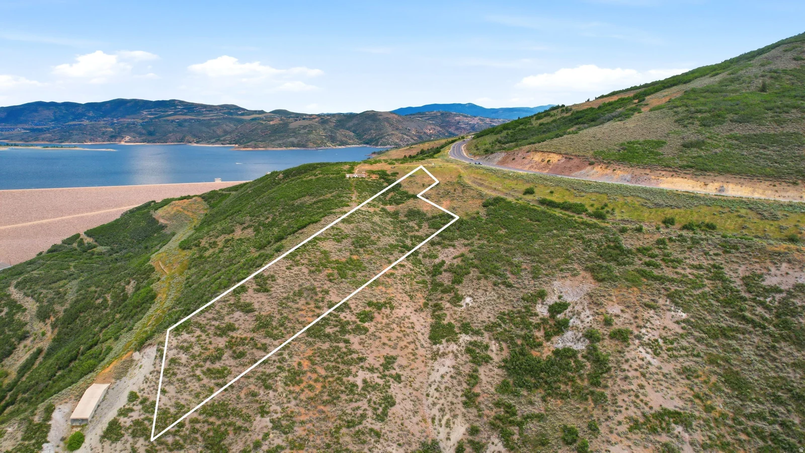 Expansive sloped land parcel featuring a visible road, surrounding hillsides, and a large body of water