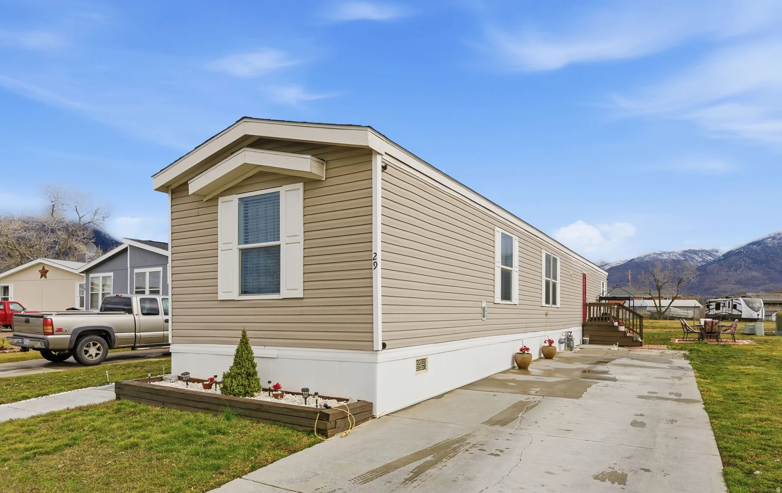 Beige vinyl siding exterior with white window shutters, a front entry deck with wood railing, a concrete driveway, and landscaped front planting bed
