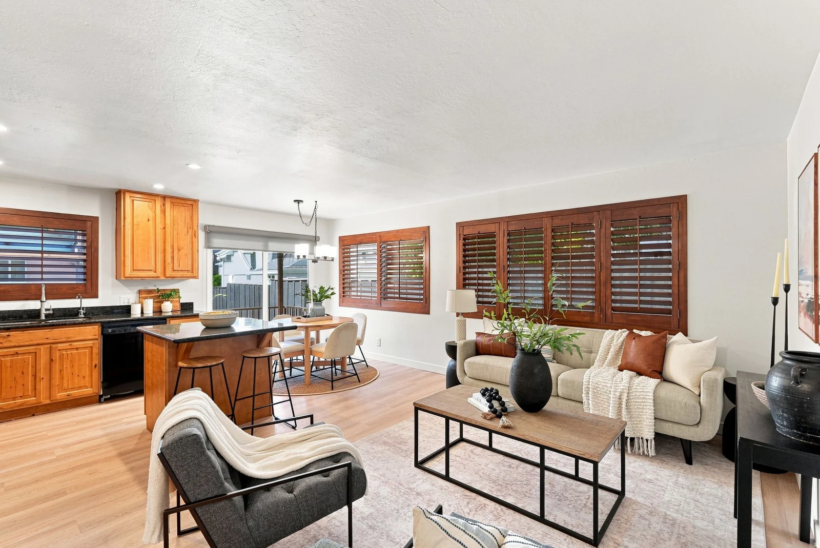 Open concept living area with wood-finish flooring, kitchen island with seating, dark countertops, and wooden plantation shutters